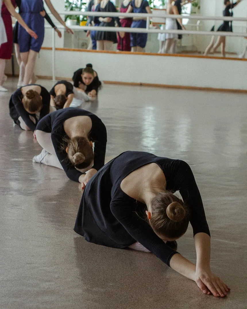 Varias bailarinas alineadas en el suelo de la sala realizando una flexión hacia delante durante el calentamiento, con la barra reflejada en el espejo