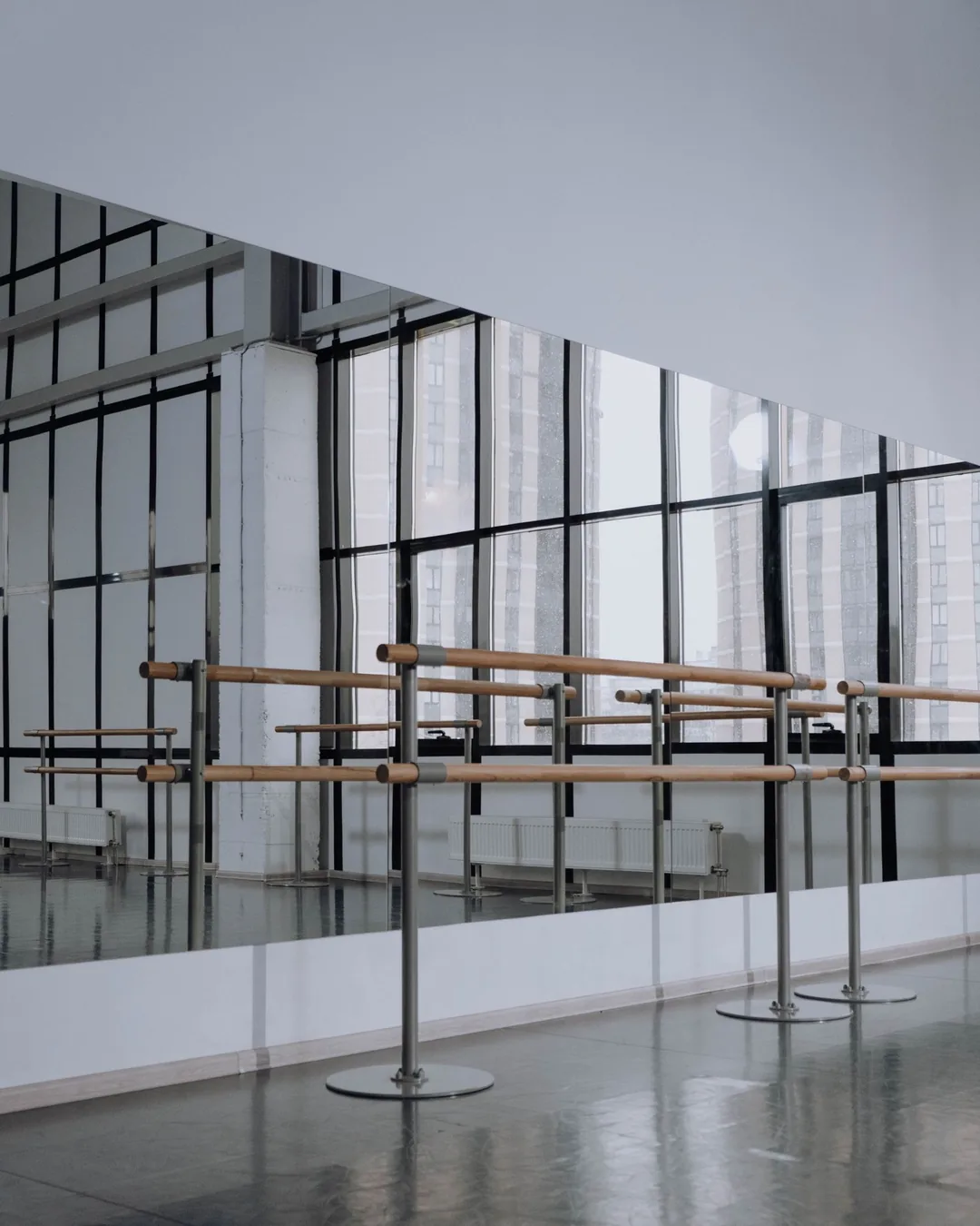 Sala de ensayo vacía con pared de espejos, barras de madera y suelo continuo iluminado por ventanales